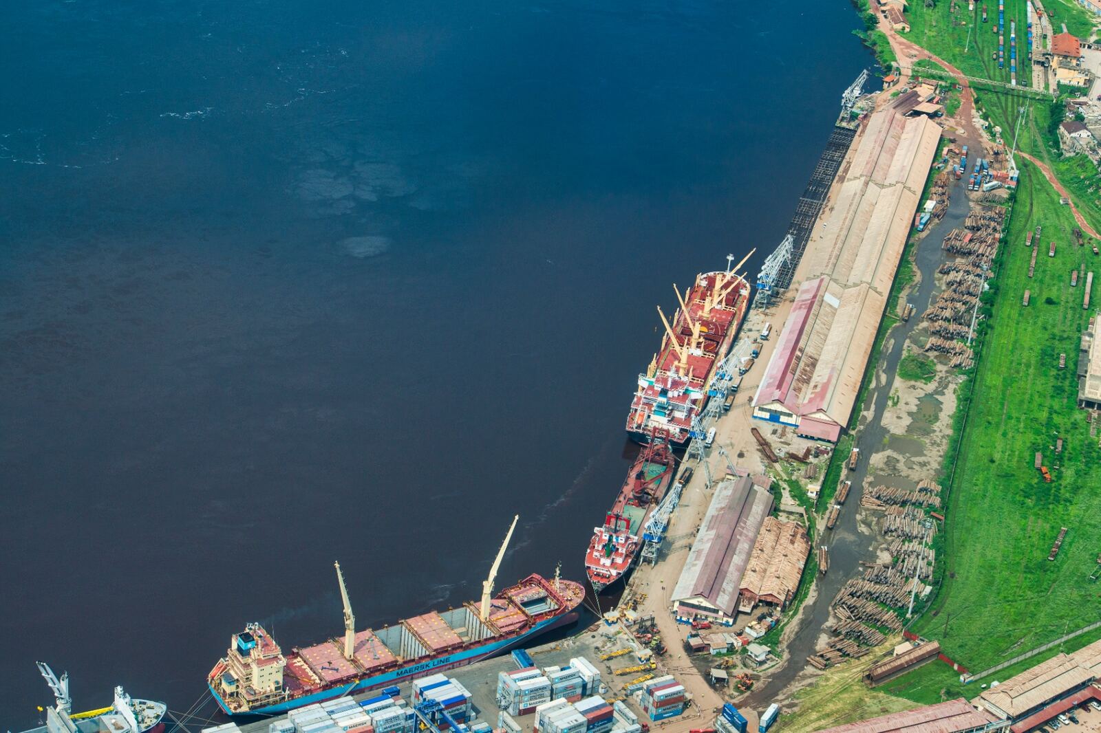 Aerial view of a river port with cargo vessels, containers and stockpiled timber alongside warehouses